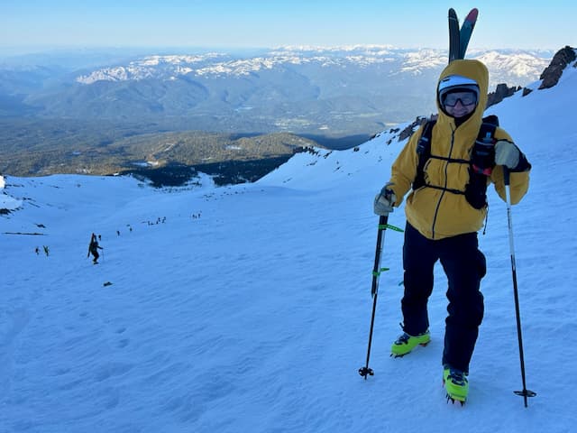 Kevin standing on the steep slope in yellow jacket with skis on pack and crampons, golden sunrise light, sweeping valley and mountain views below