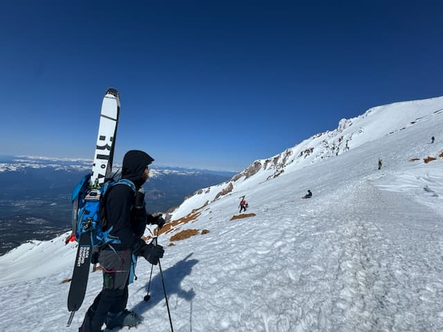 Graham pausing on the climb with skis strapped to his blue pack, looking up the route at other climbers ahead on the snowfield